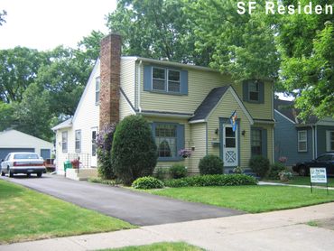A yellow two-story house with a brick chimney and a 'For Sale' sign on the lawn.