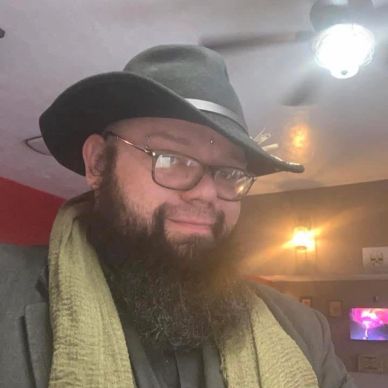 Man with glasses, a black hat, and a green scarf smiling indoors.