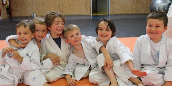 Six young boys in white martial arts uniforms sitting together on a mat, smiling.