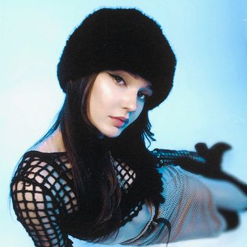 Woman in a black mesh outfit and fur hat posing against a light background.
