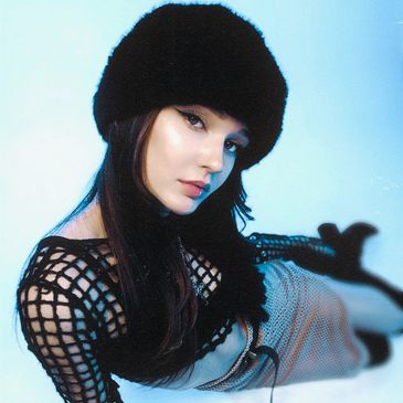 Stylish woman wearing a black fur hat and mesh top, posing with a cool blue background.