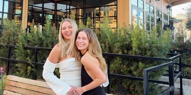 Two women smiling and posing outdoors near greenery and a modern building.
