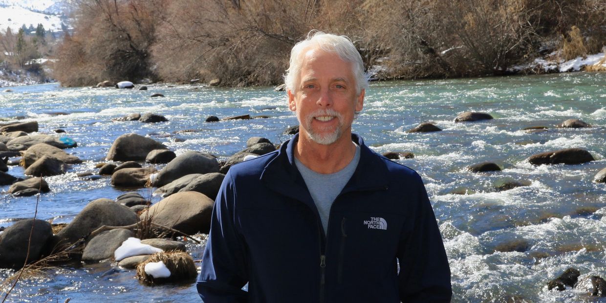 Smiling man standing by a rocky river with leafless trees in the background.
