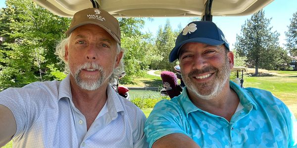 Two men smiling on a golf cart on a sunny day.