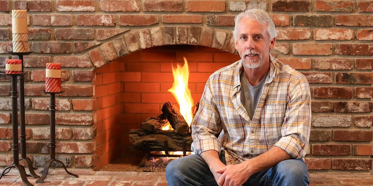 Man sitting by a cozy brick fireplace with lit candles nearby.