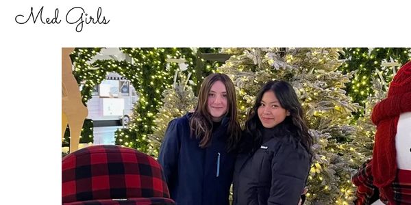 Two women posing outdoors by decorated Christmas trees with lights.