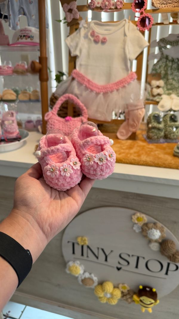 Hand holding pink crocheted baby shoes with a baby dress in the background.
