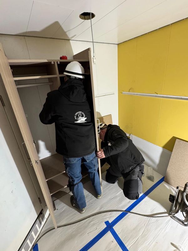 Two workers in safety gear assembling a wooden cabinet indoors.