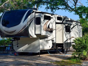 Large white fifth-wheel RV parked under trees in a sunny campground.