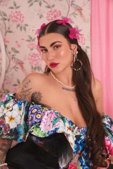 Woman with long dark hair and floral dress, adorned with pink flowers in her hair and bold makeup.