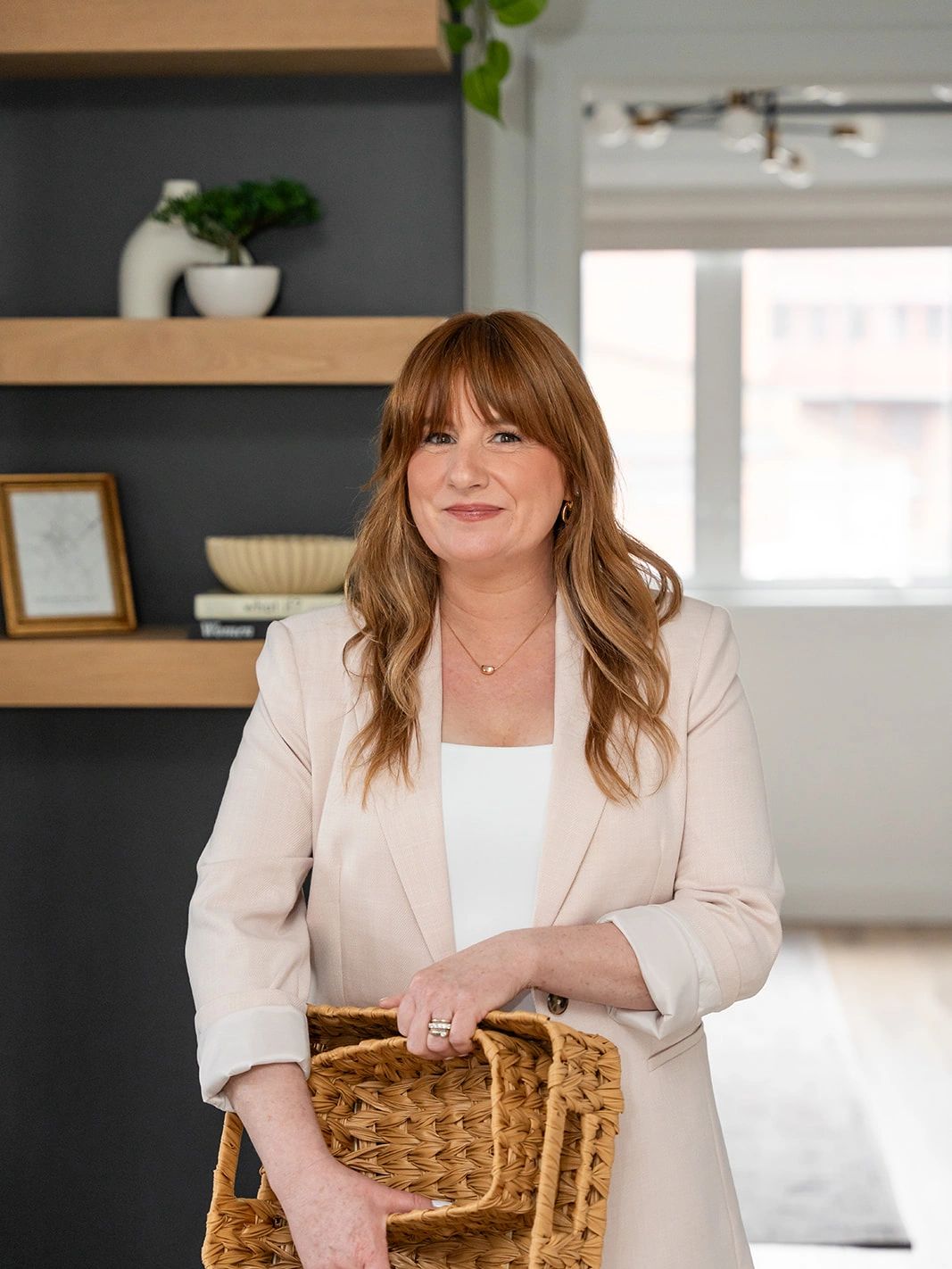 Smiling woman in a beige blazer holding a woven basket indoors.
