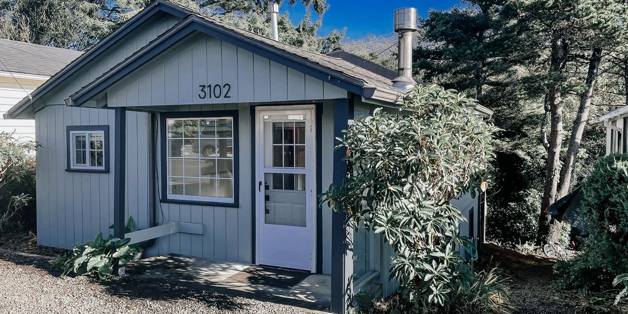 Small blue-gray cabin with white door and house number 3102 in a wooded area.