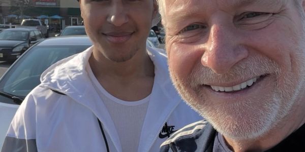 Two men smiling for a selfie outdoors near parked cars.