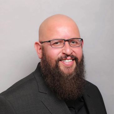 Smiling bald man with glasses and beard wearing a black suit.