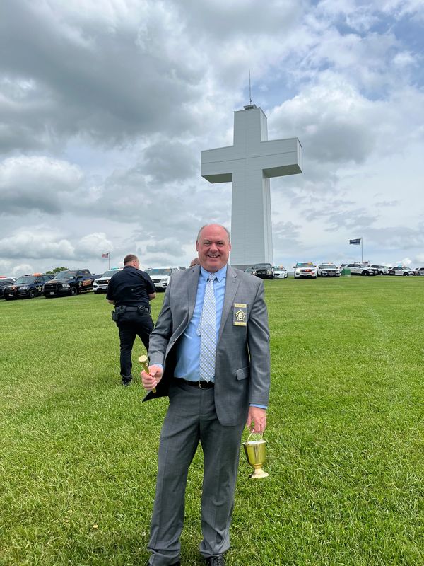 Man in suit holding a bell and trophy in front of a large white cross.