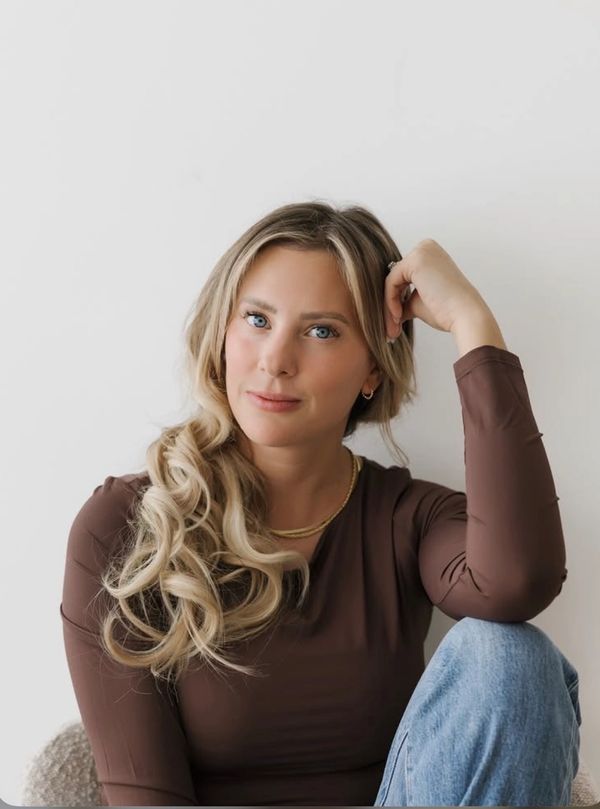 Young woman with blonde curls and blue eyes wearing a brown top and jeans.