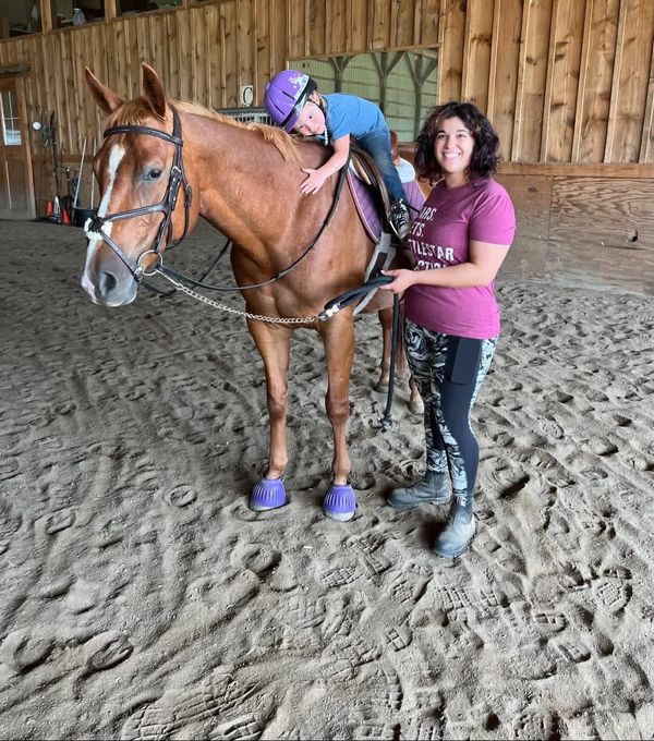 A child hugs a horse while a smiling woman stands beside them in a stable.