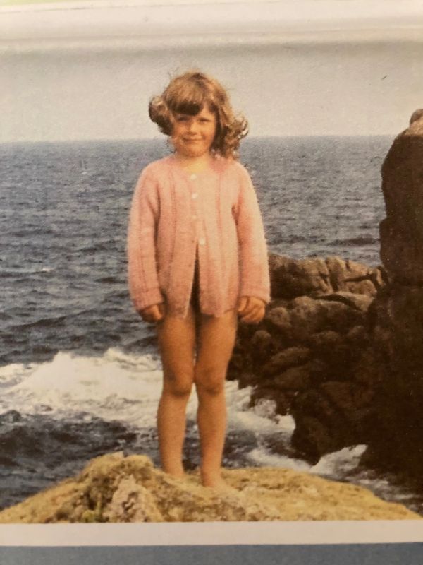 A young girl in a pink cardigan stands on a rocky shore by the sea.