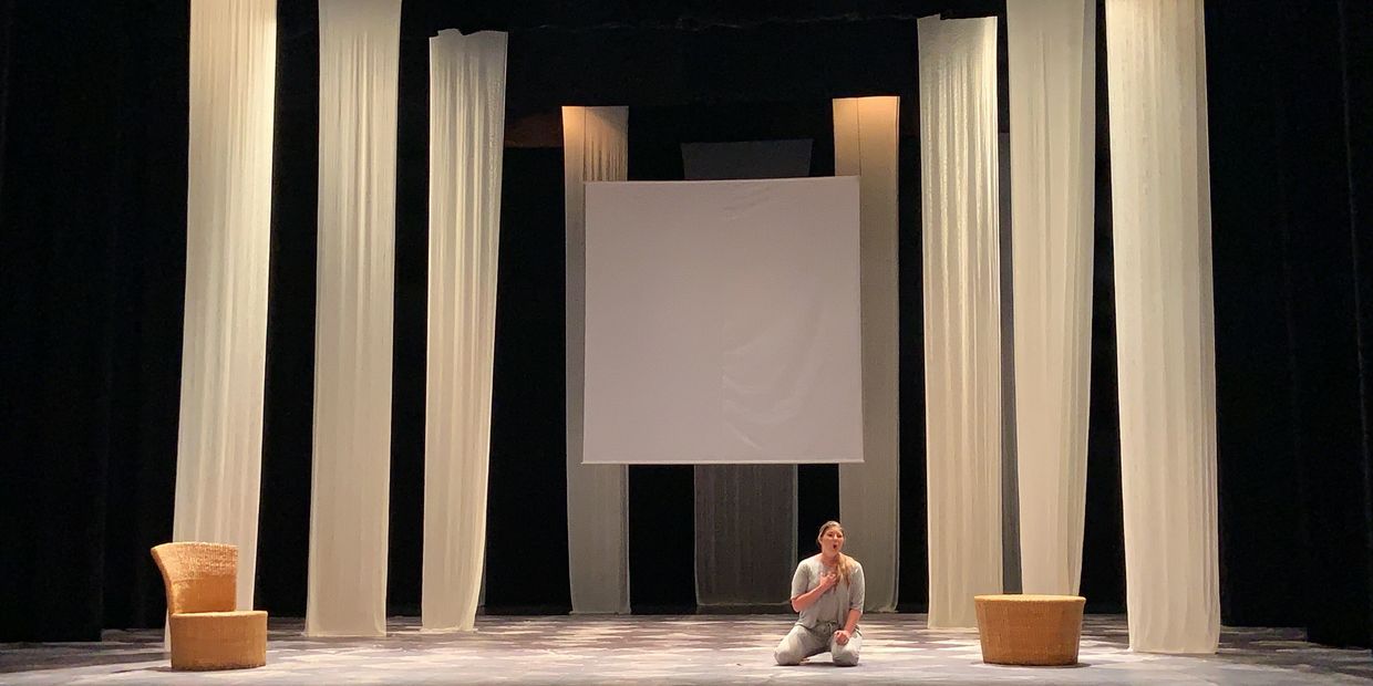 A person kneels on a minimalist stage with white curtains and wicker furniture.