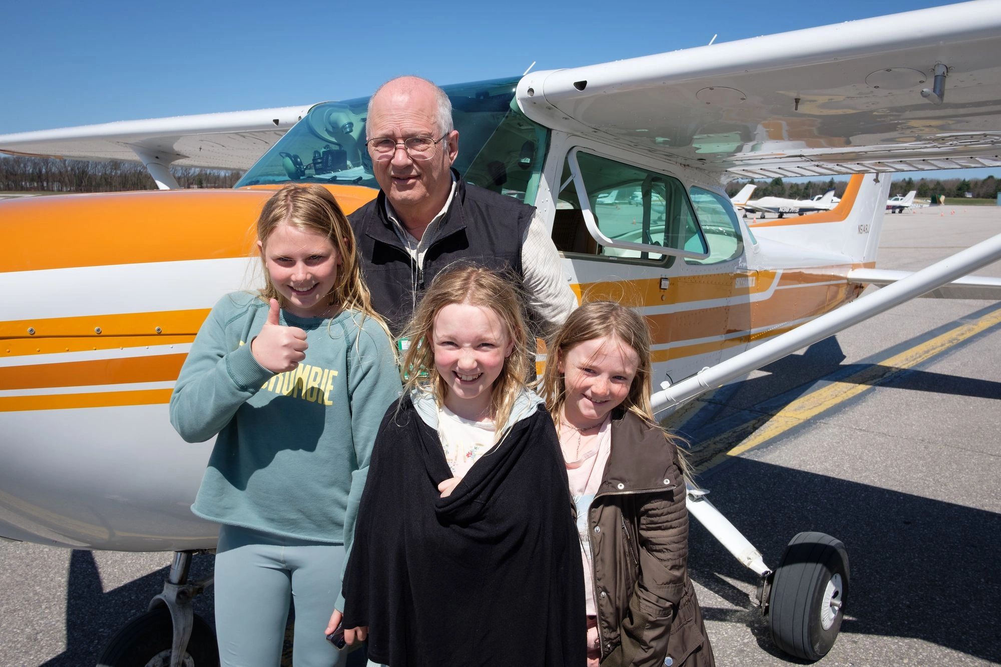 Group of three girls and an older man smiling in front of a small airplane.