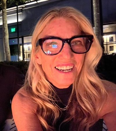 Smiling woman with long blonde hair and large glasses in a nighttime setting.