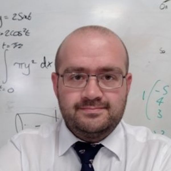 Man wearing glasses and formal attire in front of a whiteboard with equations.