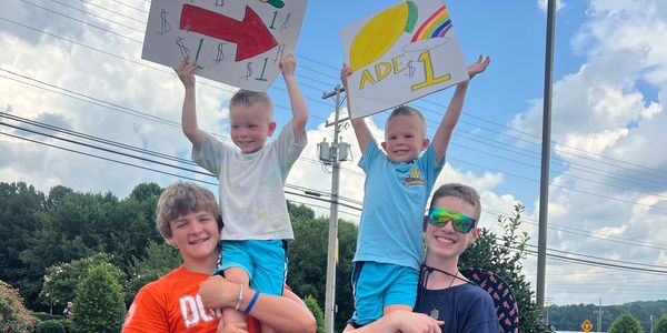 Four boys, two holding lemonade signs, smiling outdoors under a blue sky.