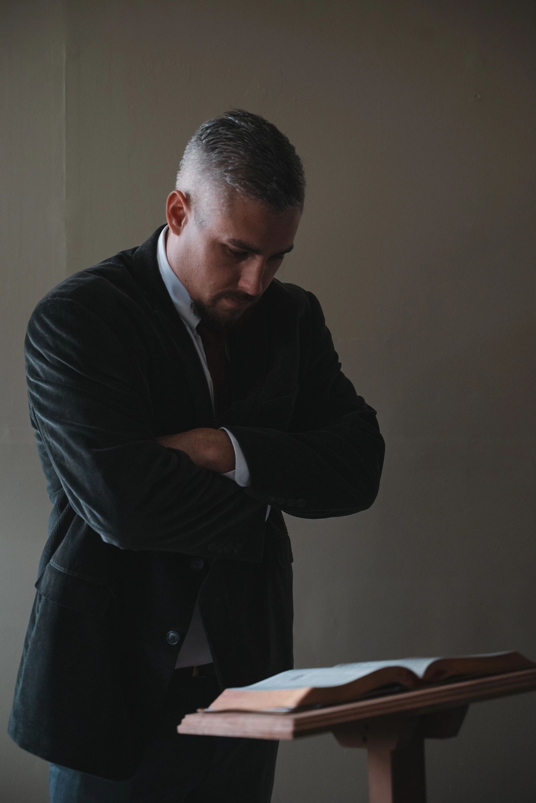 Man in a suit reading a book thoughtfully in a dim room.