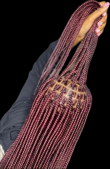 Hand holding long, neatly parted burgundy box braids against a black background.