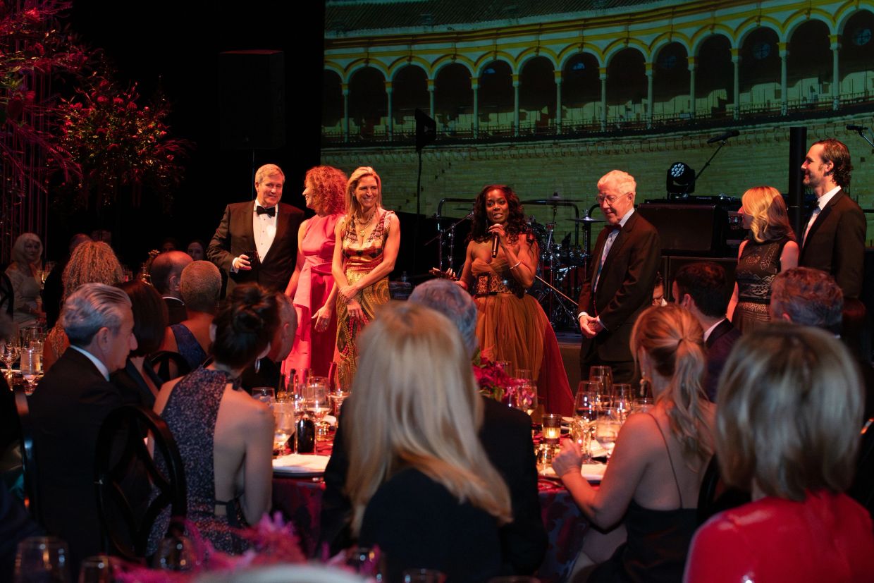 People dressed formally at an event, listening to a speaker on stage.