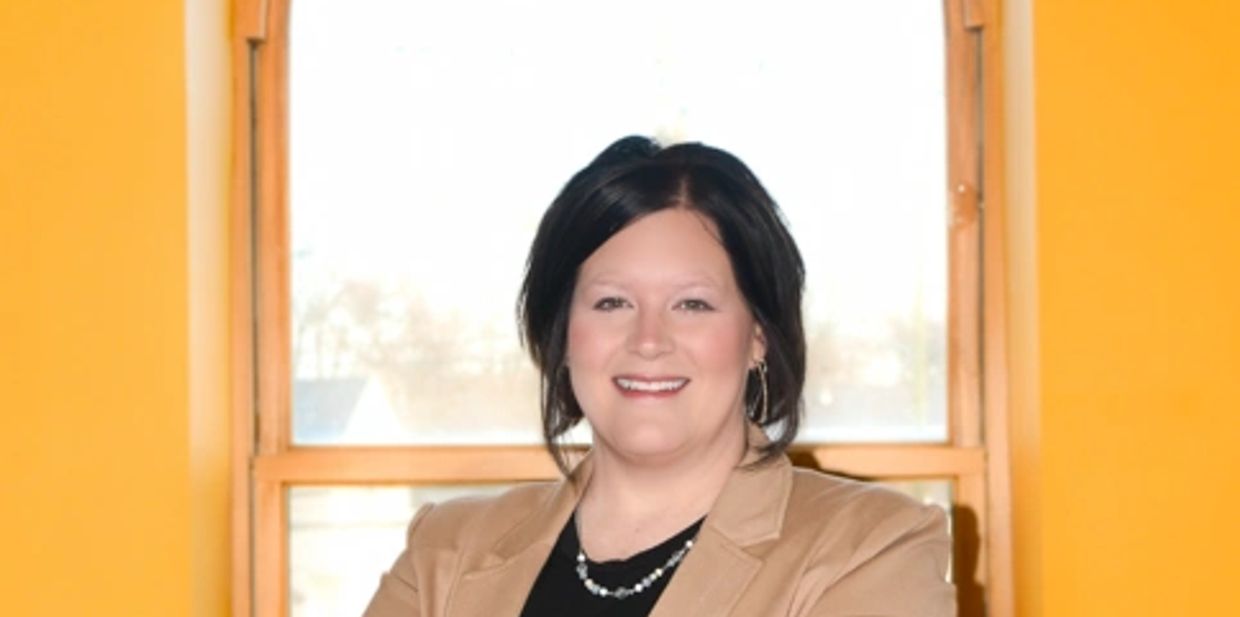 Smiling woman with dark hair wearing a tan blazer in front of a window.