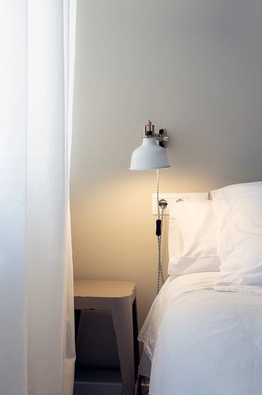 Minimalist bedroom corner with white bedding, wall lamp, and a small stool.