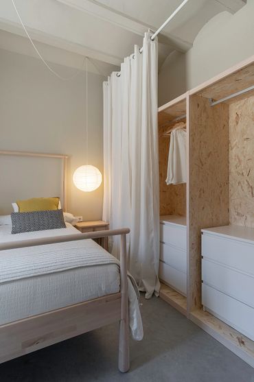 Minimalist bedroom with light wood bed, white linens, and simple wardrobe with curtain.