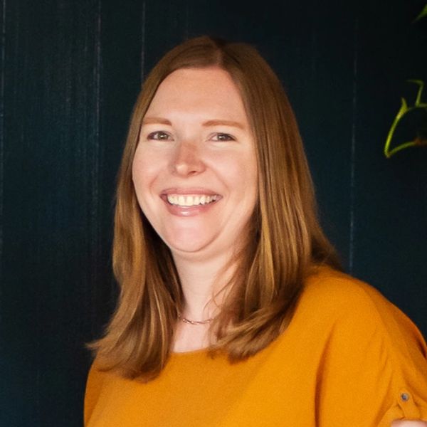 Smiling woman with long red hair in a mustard yellow top against a dark background.
