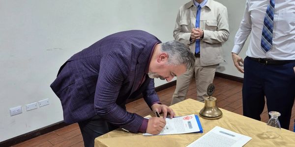 Hombre firmando documento en mesa con campana y dos personas detrás.