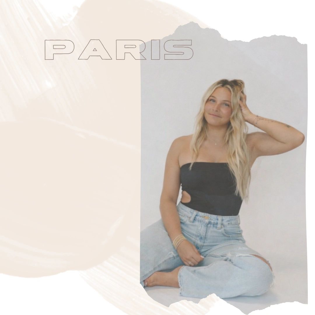 Smiling woman in black top and ripped jeans sitting against a neutral background.