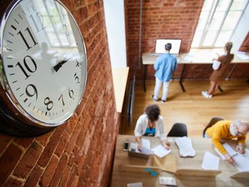 große Uhr im Vordergrund, im Hintergrund arbeitende Menschen im Coworking Space