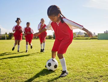 Young girl in red sports uniform controlling a soccer ball on a sunny field.