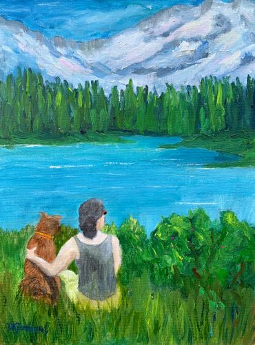 A person sits with a dog by a lake surrounded by trees and mountains.