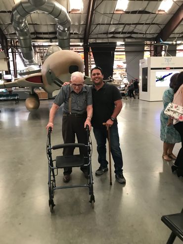 Two men posing in a large industrial setting with a vintage airplane.