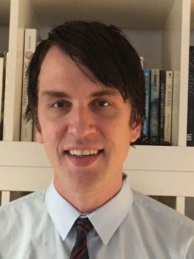 Man in a light blue shirt and striped tie smiling in front of a bookshelf.
