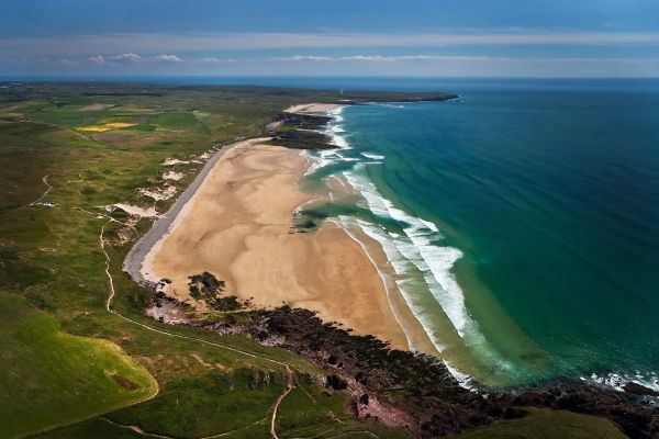 A vast sandy beach bordered by green fields and blue ocean waves under a clear sky.