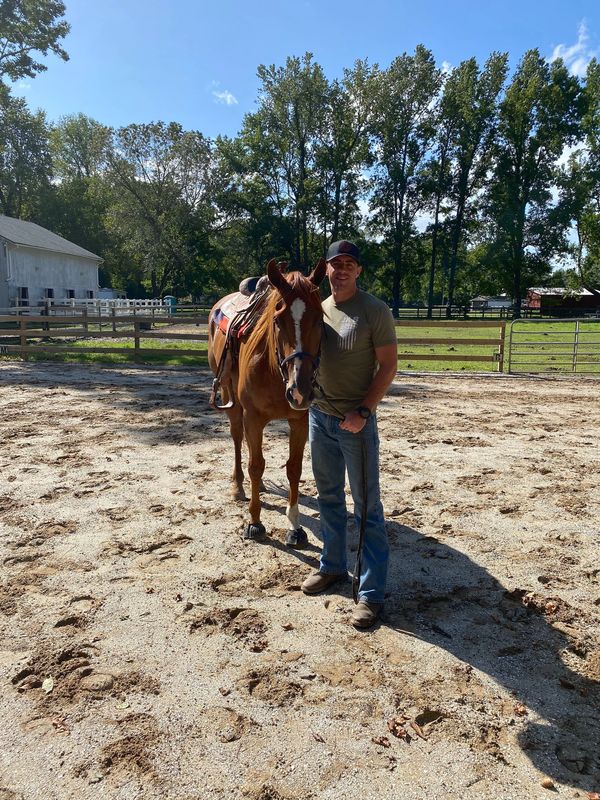 Man standing next to a saddled horse in a sunny outdoor paddock.
