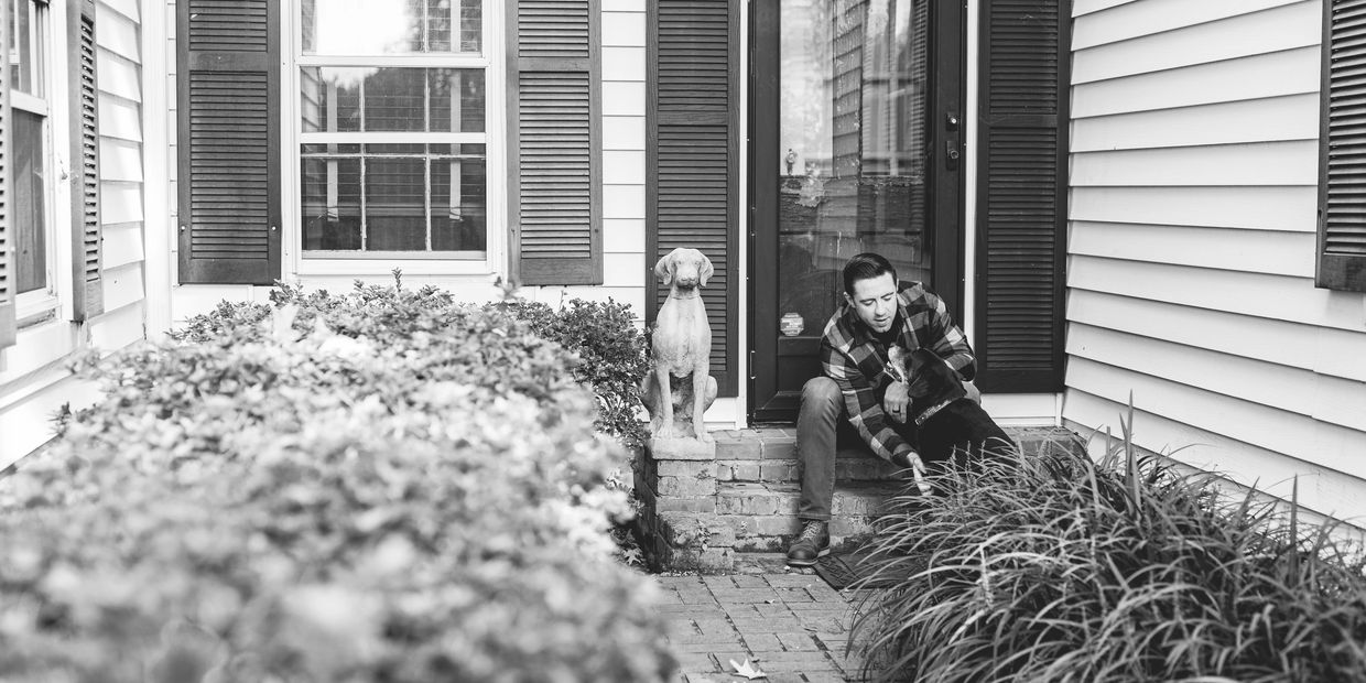 Man sitting on porch steps petting a dog beside a dog statue.