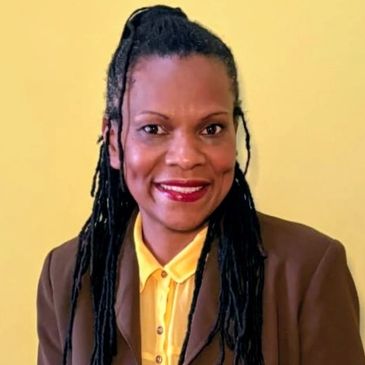 Woman with long braided hair wearing a brown blazer and yellow shirt smiling.