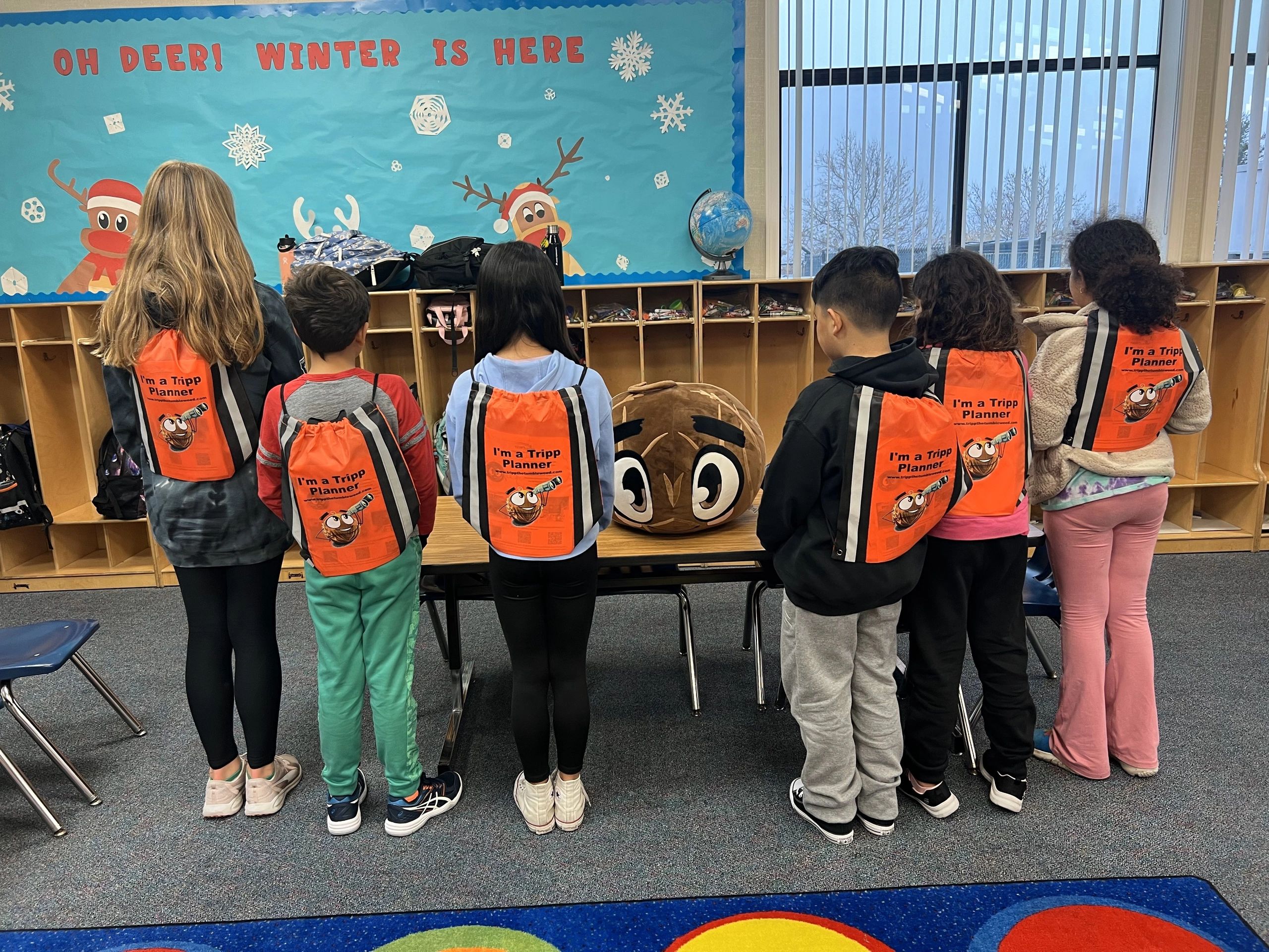 A group of students with their Tripp Planner backpacks surround a large stuffed Tripp the Tumbleweed