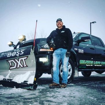 Man stands proudly by a snowplow truck labeled 'Oelkers Lawn Care' on a snowy day.