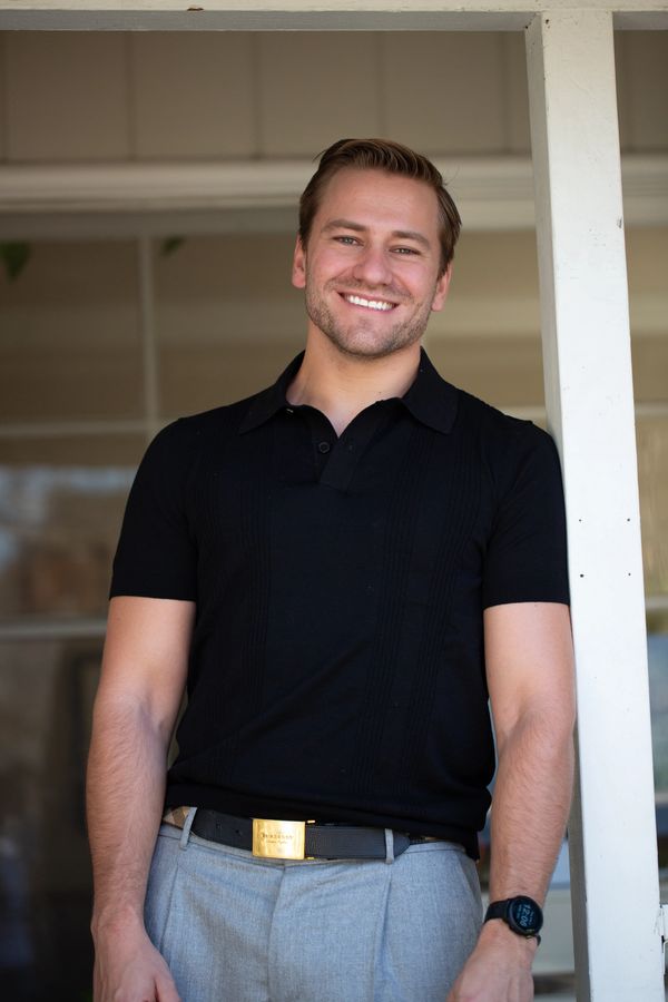 Smiling man in black shirt and gray pants standing outdoors.