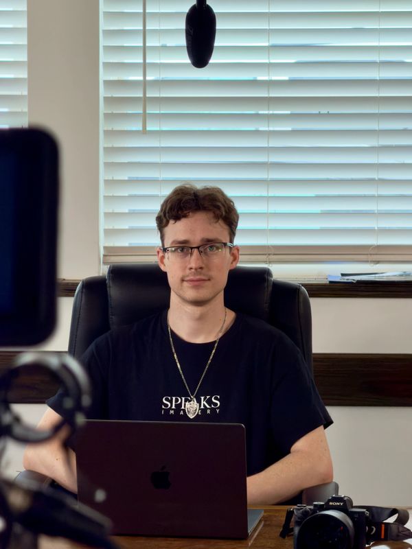 Man sitting at desk with laptop and camera, facing camera.