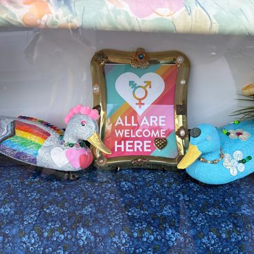 Decorative ducks flank a framed sign reading 'All Are Welcome Here' with a gender-inclusive symbol.